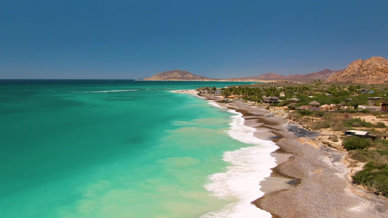 Aerial frame of Cabo Pulmo coastline with pale turquoise shallows, a bright band of foam on the beach, scattered small buildings along the vegetation line, arid hills beyond, and no visible people.
