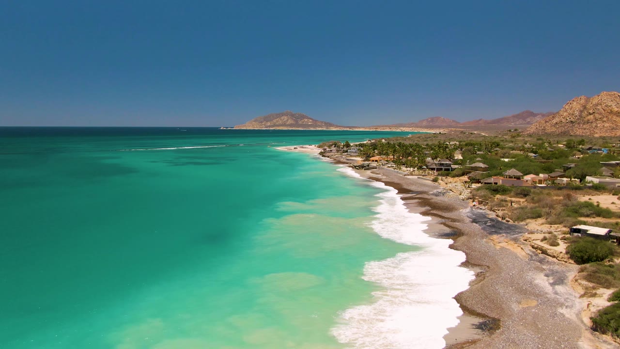 Cabo Pulmo aerial view with a wider stretch of turquoise ocean, white waves curling onto the shore, small roofed structures set back from the beach, rugged hills in the background, and no visible people.