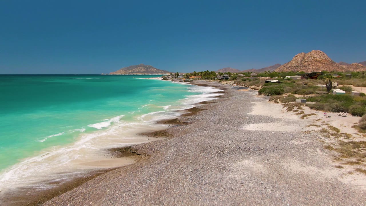 Aerial frame over Cabo Pulmo with small white waves touching the pebbled shore, pale sand patches along the beach, low scrub inland, distant hills, and no visible people.