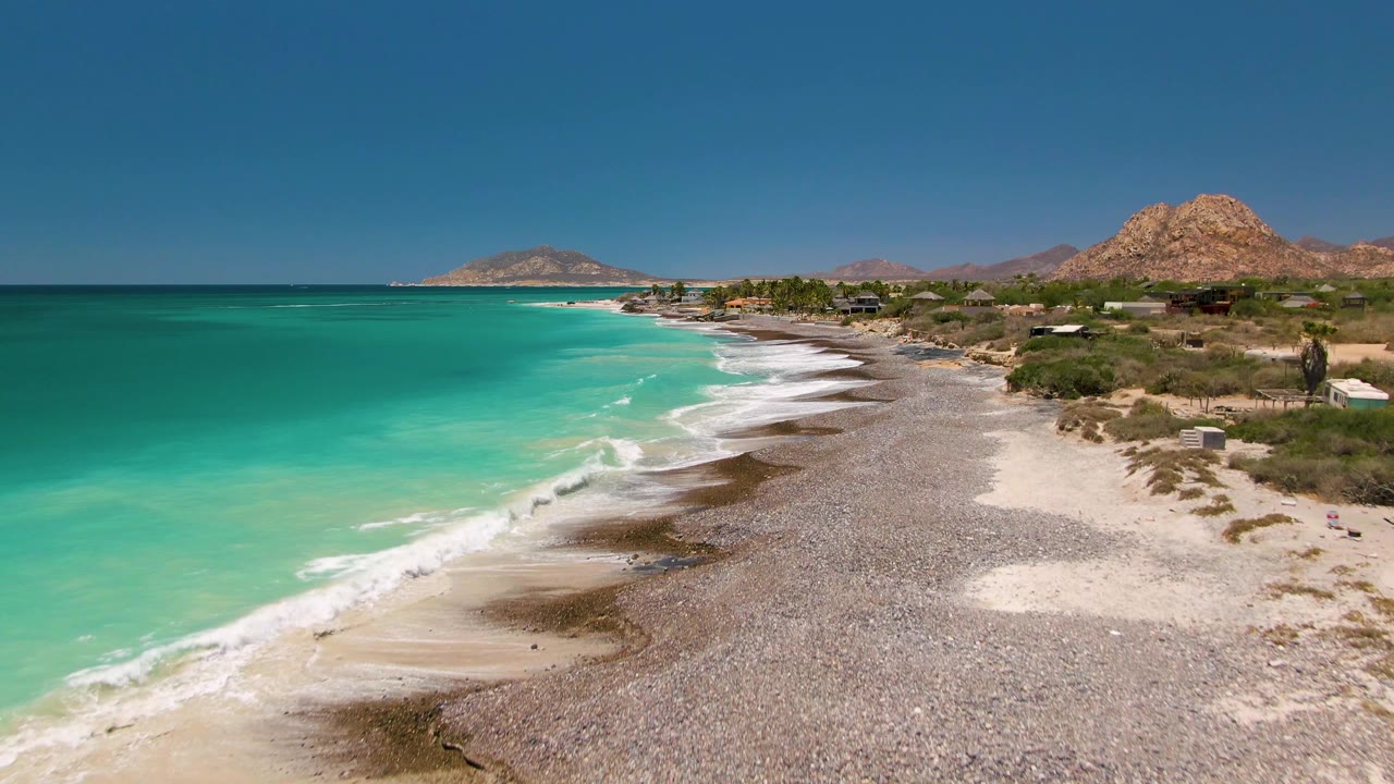 Aerial frame over Cabo Pulmo showing the beach curve extending toward distant hills, turquoise water filling the left side, scrubby coastal land on the right, and no visible people.