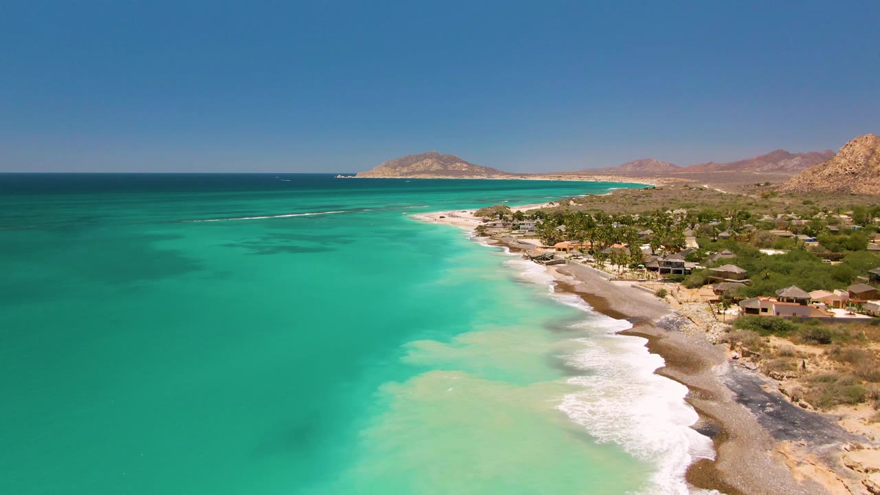 Aerial frame over Cabo Pulmo showing turquoise water, a curved sandy shoreline, scattered coastal buildings, arid hills, clear blue sky, and no visible people.