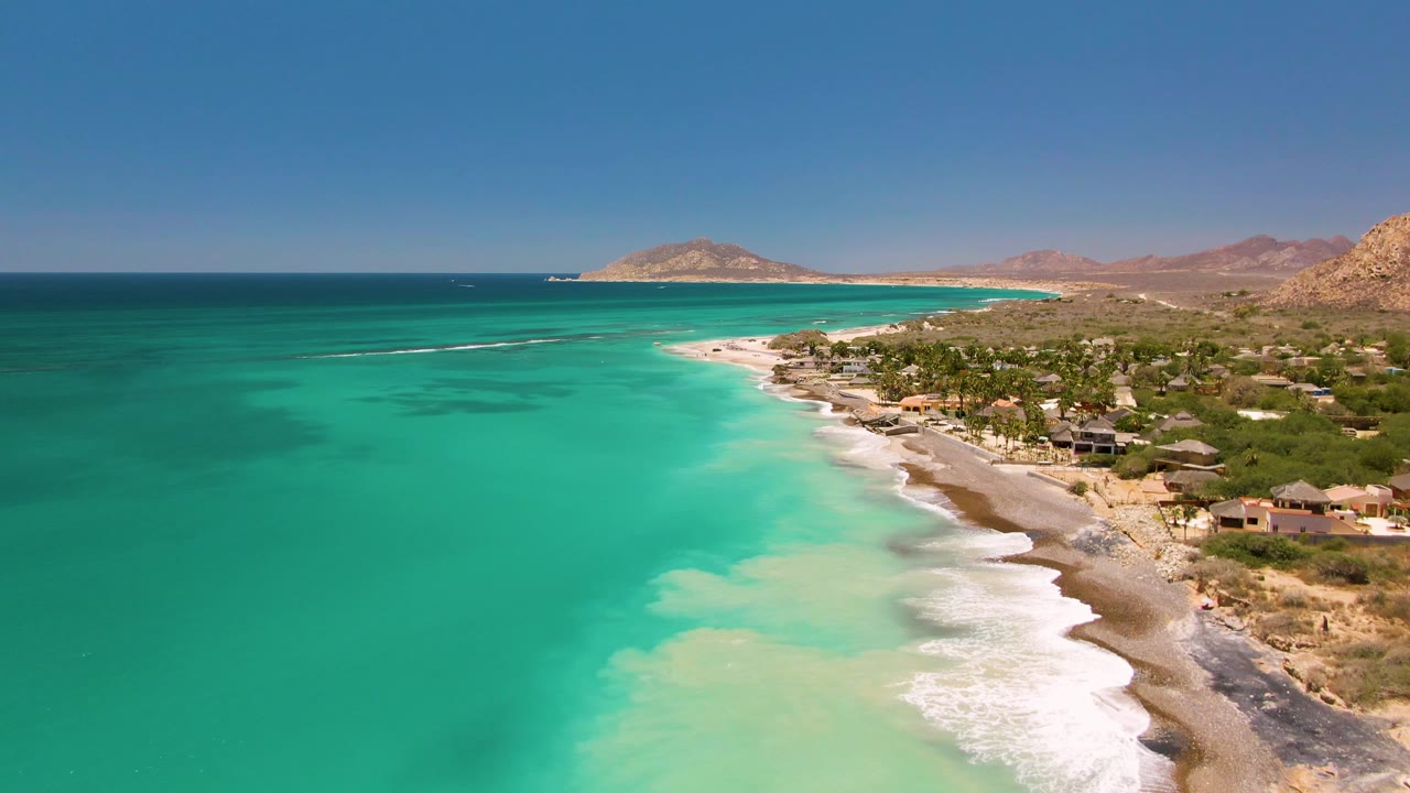 Turquoise water and white surf along a sandy shoreline beside greenery and low coastal buildings.