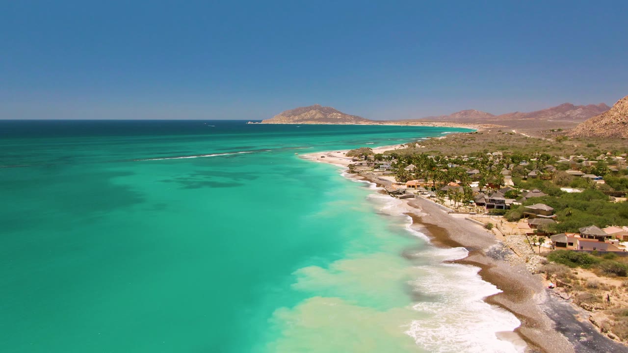 Cabo Pulmo coastline seen from above with broad turquoise ocean on the left, a foamy beach edge, coastal homes and trees on the right, dry hills beyond, and no visible people.