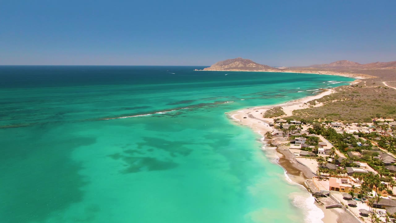 At 0:00, Cabo Pulmo shoreline appears from above with turquoise water in the foreground, a sandy beach curving along the right, low coastal buildings, distant tan hills, and no visible people.