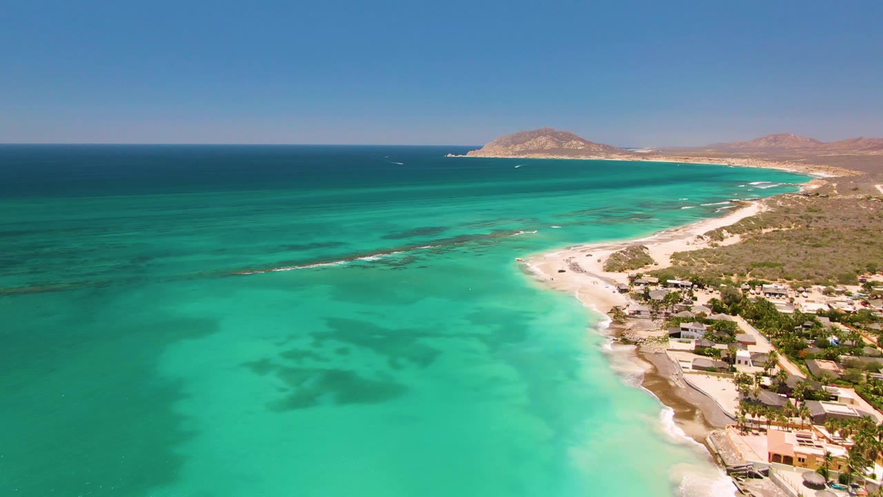 Wide drone view of clear turquoise water, white surf, beach, buildings, and hills.