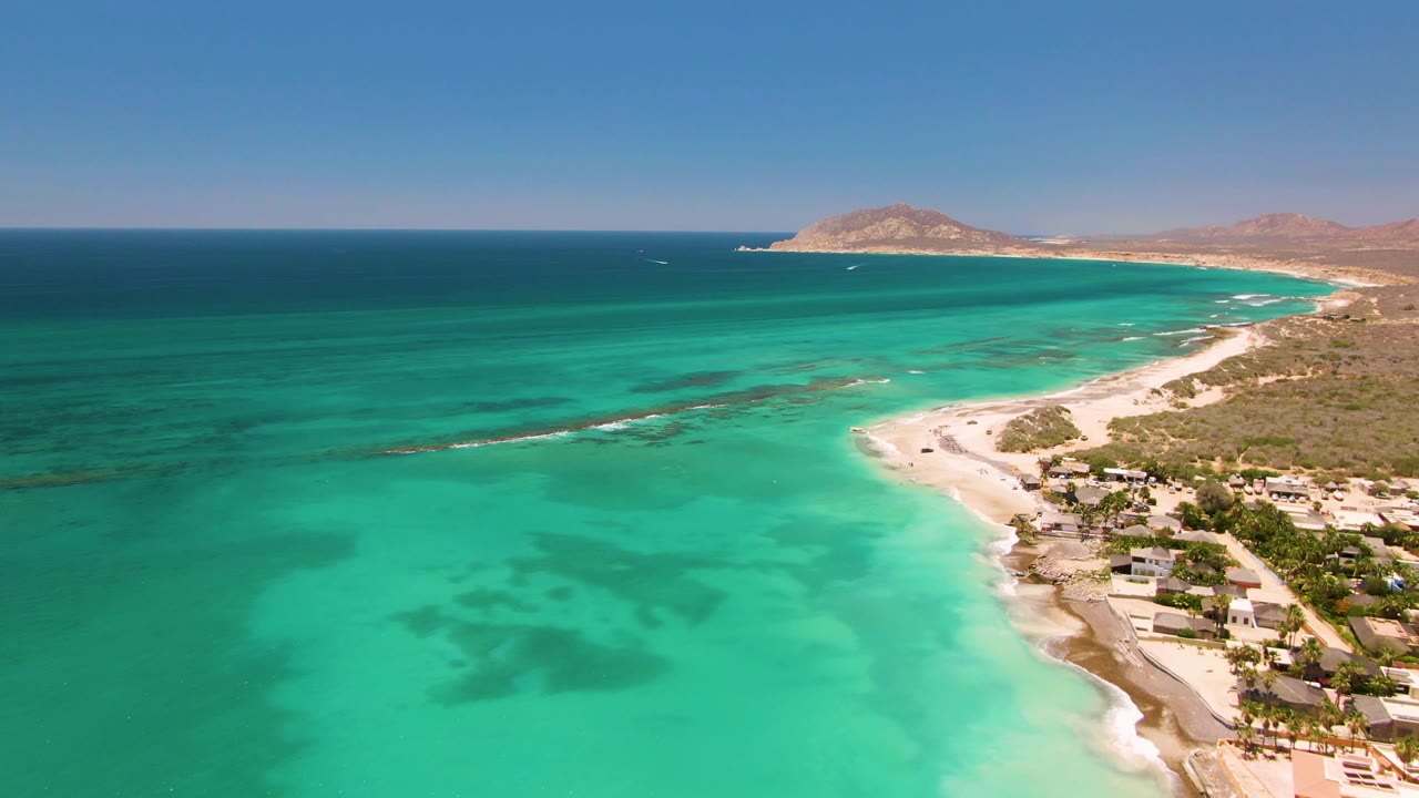 At 0:02, the Cabo Pulmo coast remains in a wide aerial composition with teal water dominating the frame, the beach and buildings tight along the right side, distant hills, and no visible people.
