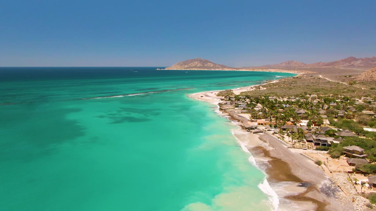 Aerial view over Cabo Pulmo turquoise water, sandy beach, shoreline buildings, palm trees, arid hills, and no visible people at the start of the clip.