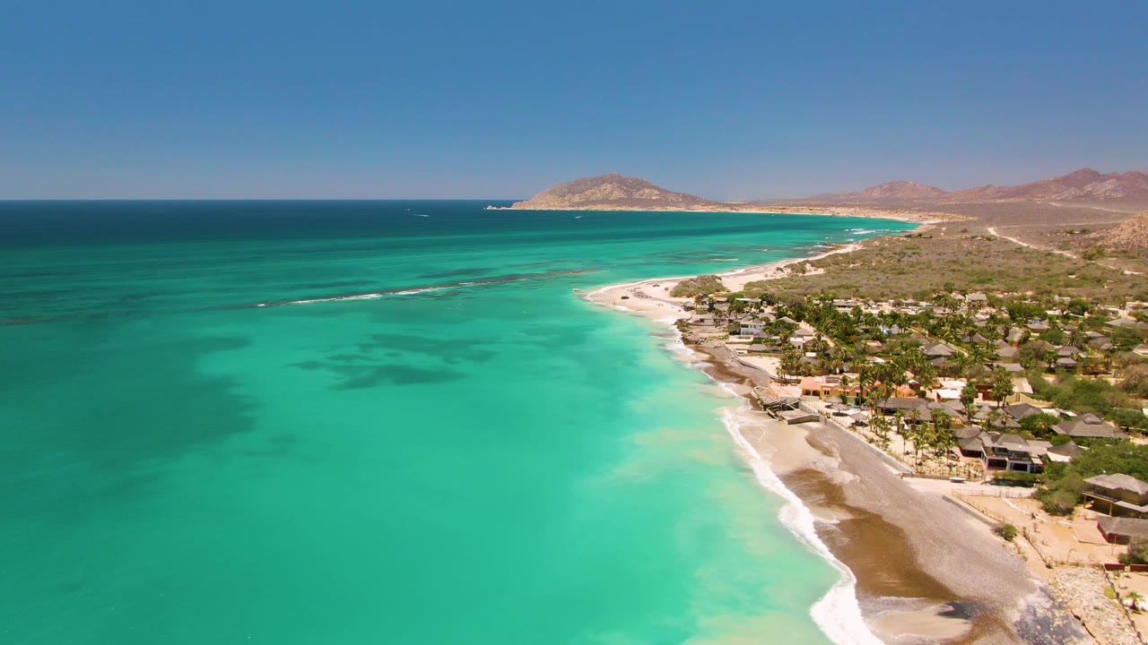 Turquoise water and white surf line a sandy beach with buildings inland.