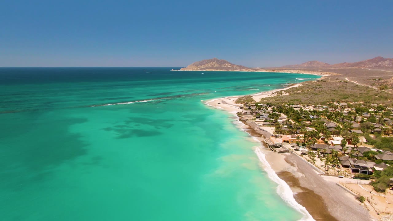 Wide Cabo Pulmo aerial with open turquoise shallows, a sandy curve of beach, low coastal development at right, desert hills in the distance, and no visible people.