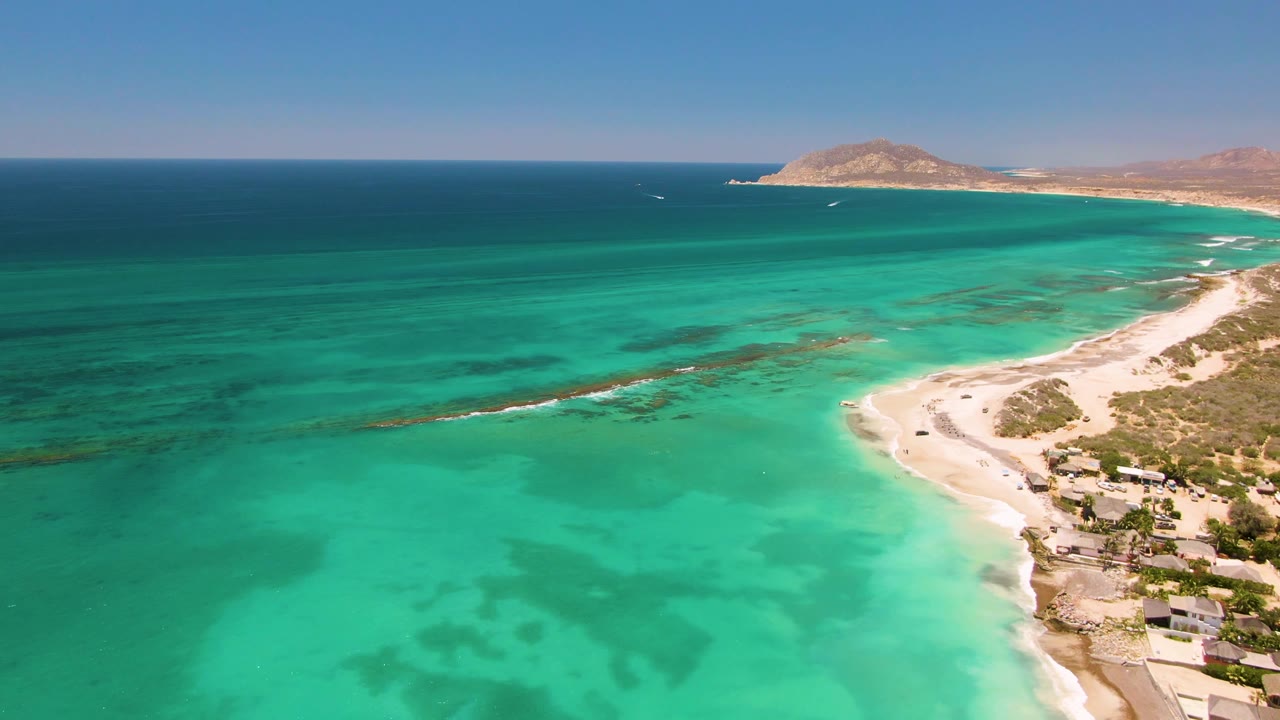 Aerial frame over Cabo Pulmo, Mexico, with turquoise shallows in the foreground, a curving sandy beach at right, distant dry hills, and no visible people.
