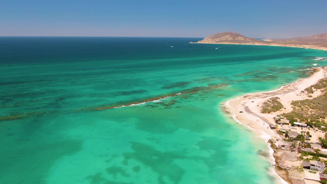 Drone view over Cabo Pulmo with shallow turquoise water covering most of the frame, pale beach on the right, small coastal structures near shore, and no visible people.