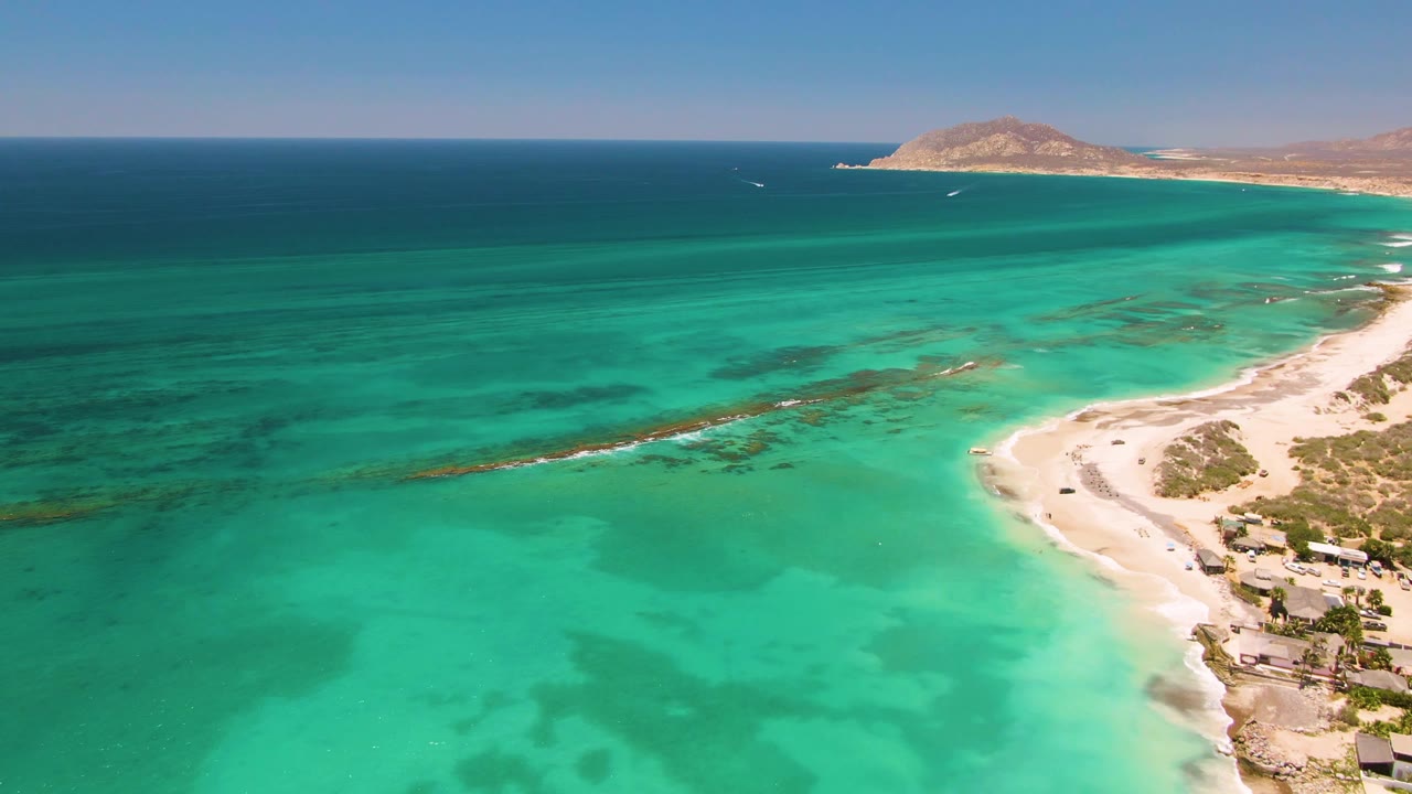 Wide aerial frame of Cabo Pulmo showing turquoise sea across the foreground and midground, a sandy point along the right side, low structures near shore, and no visible people.