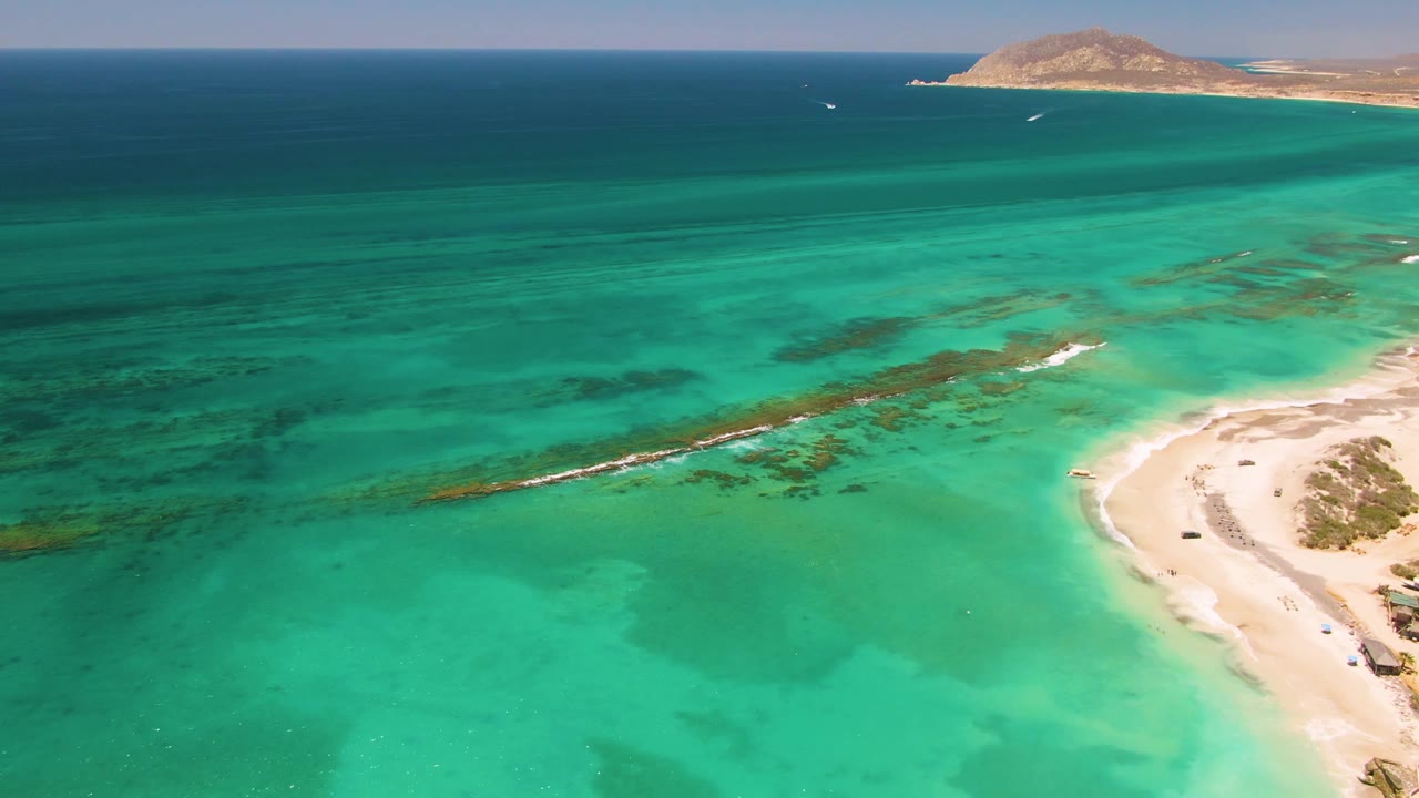 Aerial view over Cabo Pulmo turquoise shallows with a pale sand beach on the right, darker seabed patches across the water, distant land on the horizon, and no visible people.