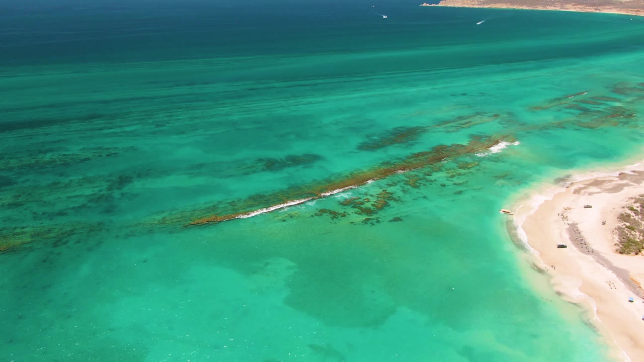 Aerial view of Cabo Pulmo coastline with broad turquoise water in the foreground, darker seabed areas through the center, a pale beach along the right, distant headland at top right, and no visible people.