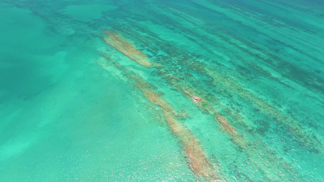 Overhead view of shallow turquoise water and diagonal coral reef bands in Cabo Pulmo, with a small reddish fish-like shape near the reef and no visible people.