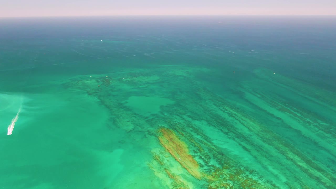 Aerial Cabo Pulmo frame with the small boat farther into the turquoise water, reef bands visible below, deeper blue water beyond, and no visible people.