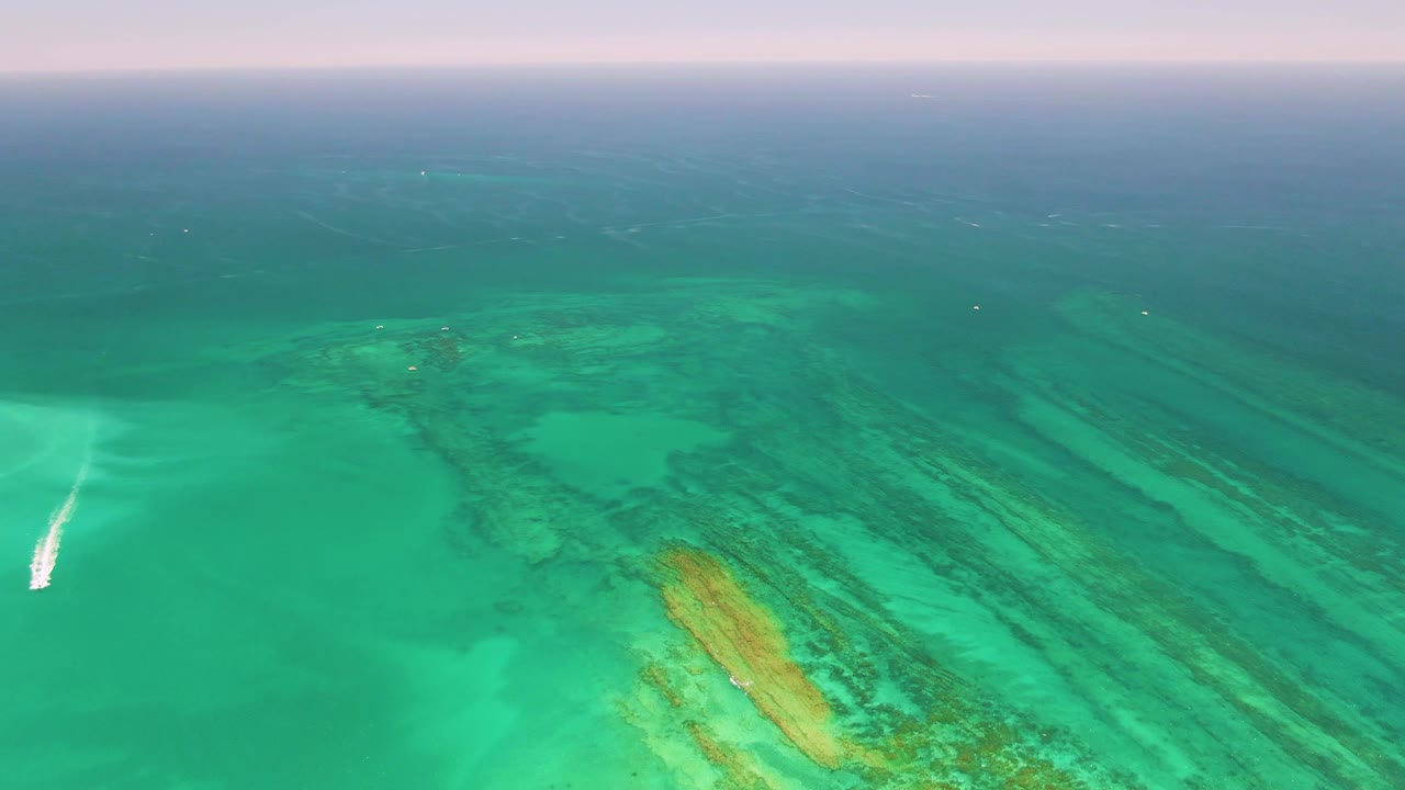 Mid-clip aerial over Cabo Pulmo with the boat partly near the left edge, a bright wake on turquoise water, layered reef forms below, and no visible people.