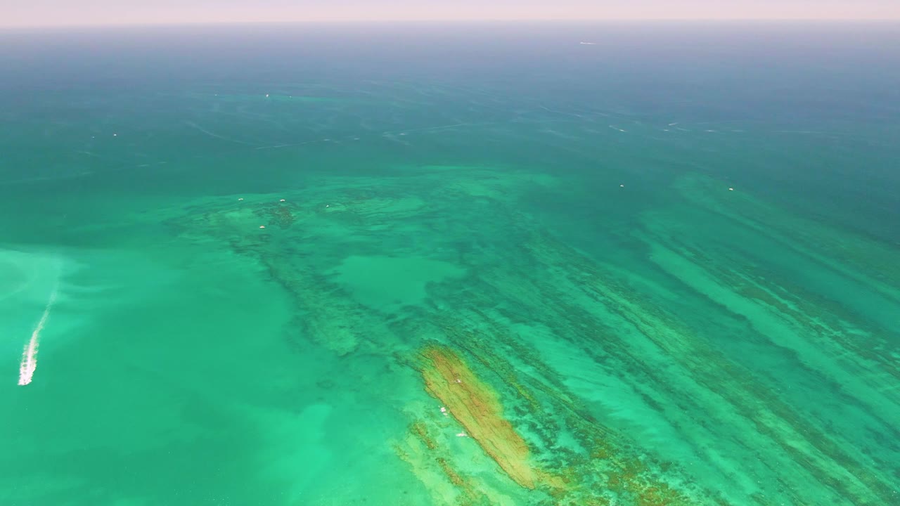 Aerial view of turquoise water over a coral reef with a white boat wake at left.
