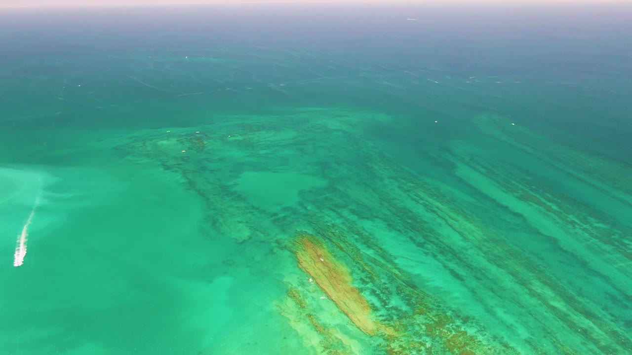 Aerial frame over Cabo Pulmo shows green-blue water with submerged reef lines, a bright white boat wake closer to center-left, a hazy horizon band, and no visible people.