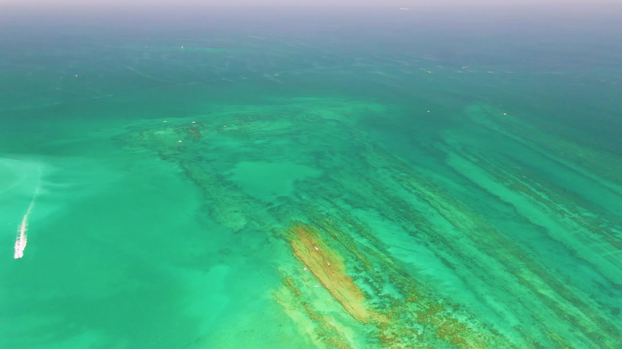 Aerial frame over Cabo Pulmo captures turquoise shallows with yellow-green reef patches, darker reef streaks, a white wake near the left side, and no visible people.