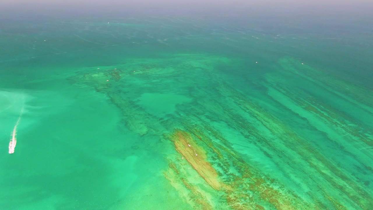 Aerial frame over Cabo Pulmo shows the wake tight to the far left edge, broad turquoise water in the center, reef texture running diagonally below, and no visible people.