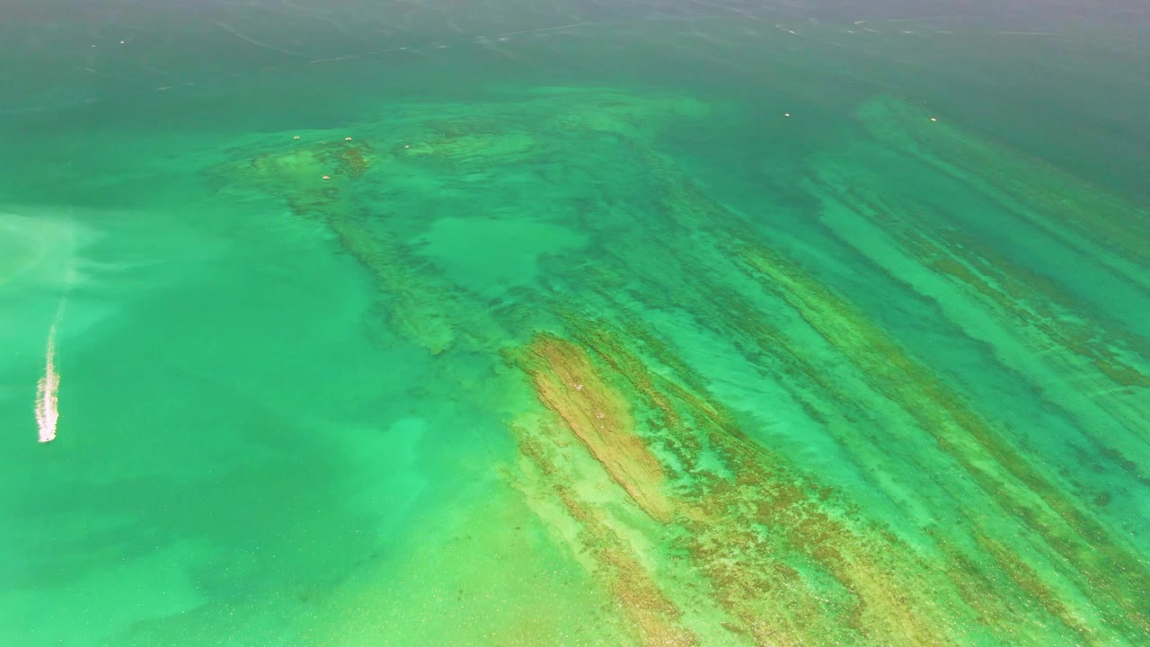 At 0:02, the small boat remains left of center in the Cabo Pulmo water, while a broad tan reef patch and darker coral streaks run diagonally across the right side, with no visible people.