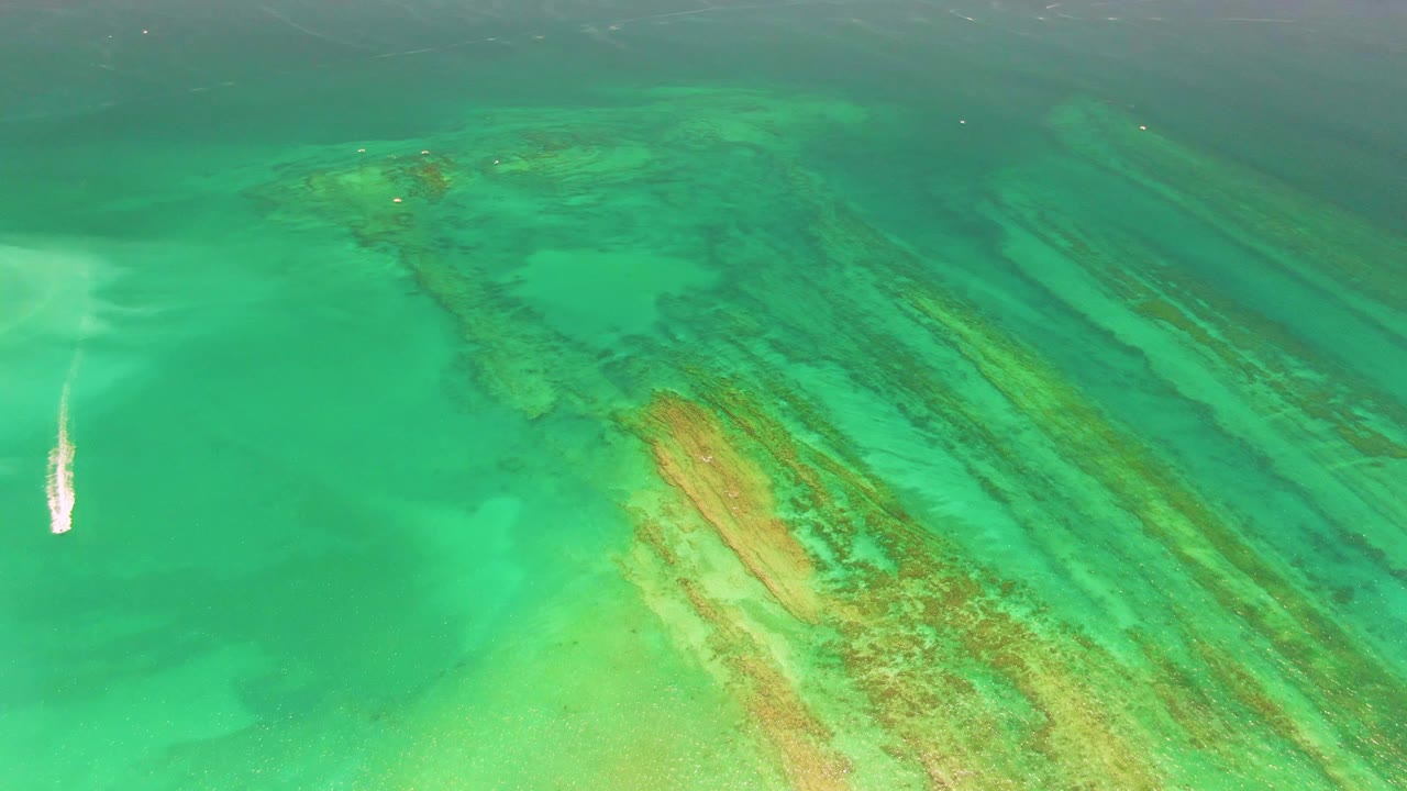 At 0:03, the boat shifts toward the lower-left edge of the Cabo Pulmo aerial frame, leaving a vertical white wake as reef bands fill the right side, with no visible people.