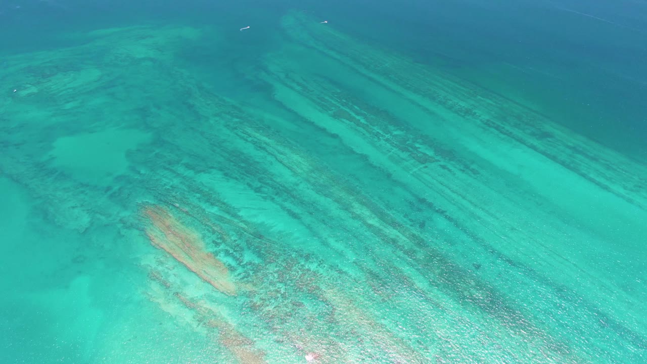 A high aerial frame over Cabo Pulmo turquoise water shows pale sandy shallows, darker reef bands, a warm coral-toned patch near the lower left, and no visible people.