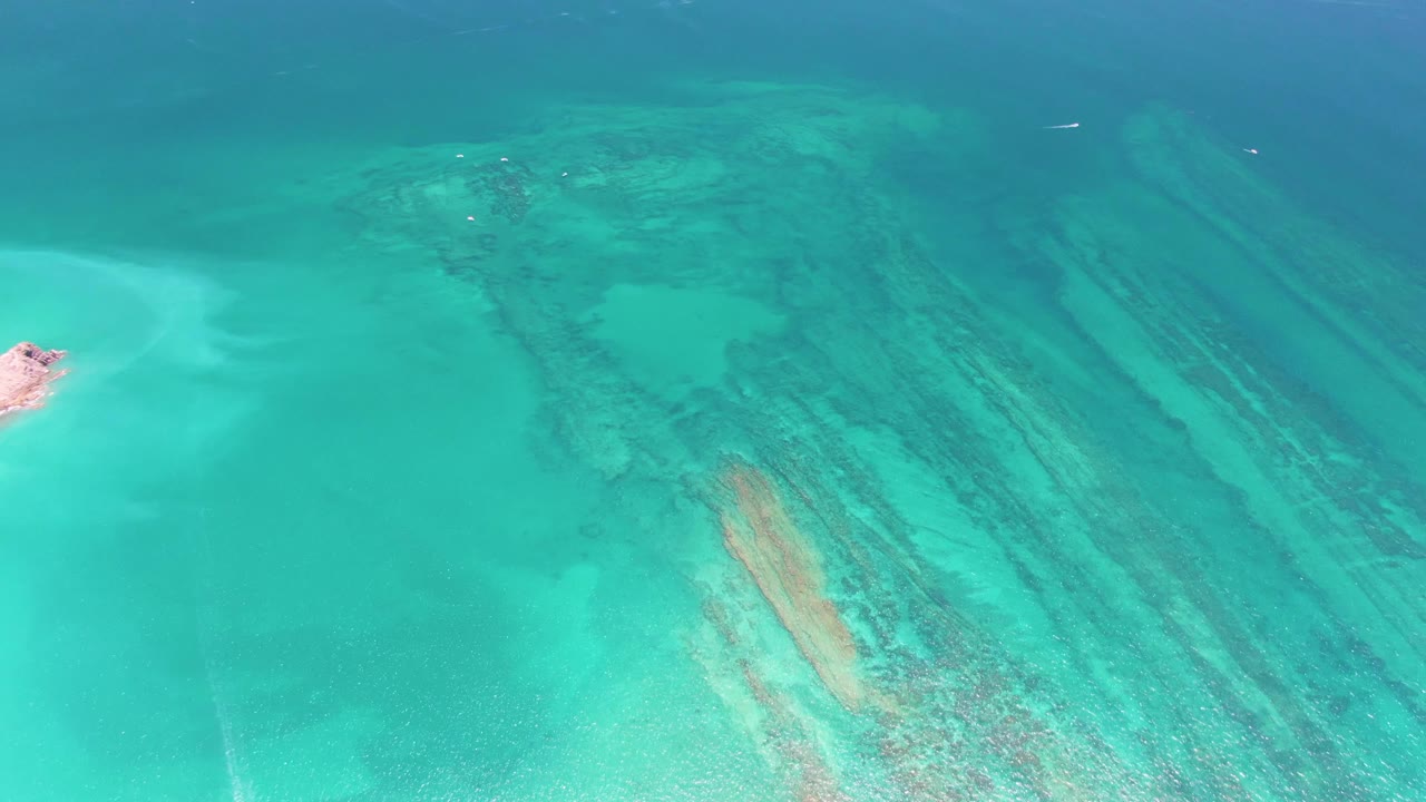 Overhead view of clear turquoise water in Cabo Pulmo with pale sand, submerged dark reef streaks, a small sandy point at the left edge, and no visible people.