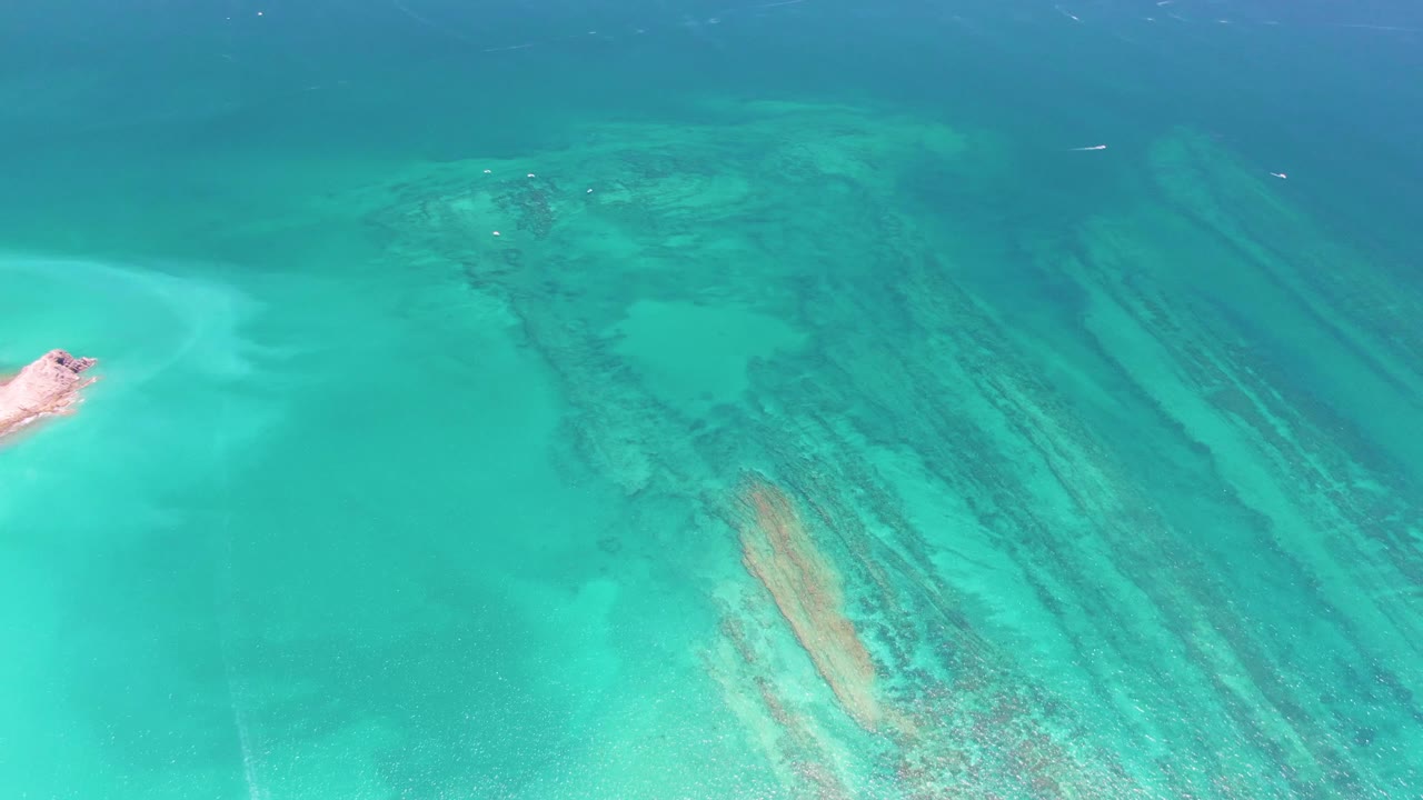 Aerial frame over Cabo Pulmo water with a pale sand patch at left, dark coral-like bands crossing the shallows, deeper blue water above, and no visible people.