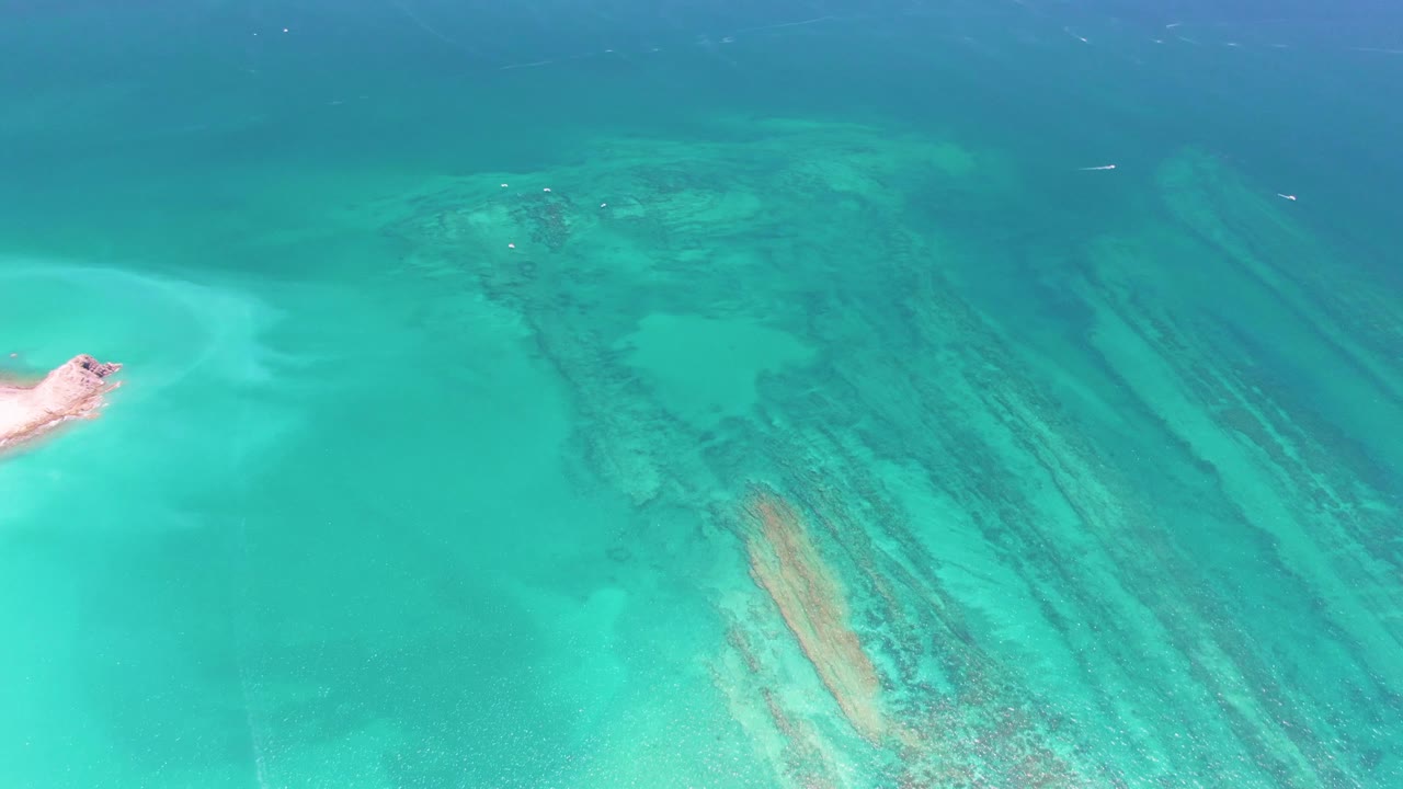 High overhead Cabo Pulmo frame with turquoise shallows, elongated dark reef marks, a pale sand shape near the left side, light surface texture, and no visible people.