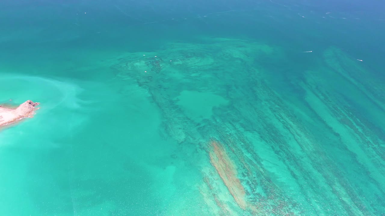 Overhead view in Cabo Pulmo showing a wider spread of turquoise shallows, dark reef formations below the surface, a sandy point clipped at the left edge, and no visible people.