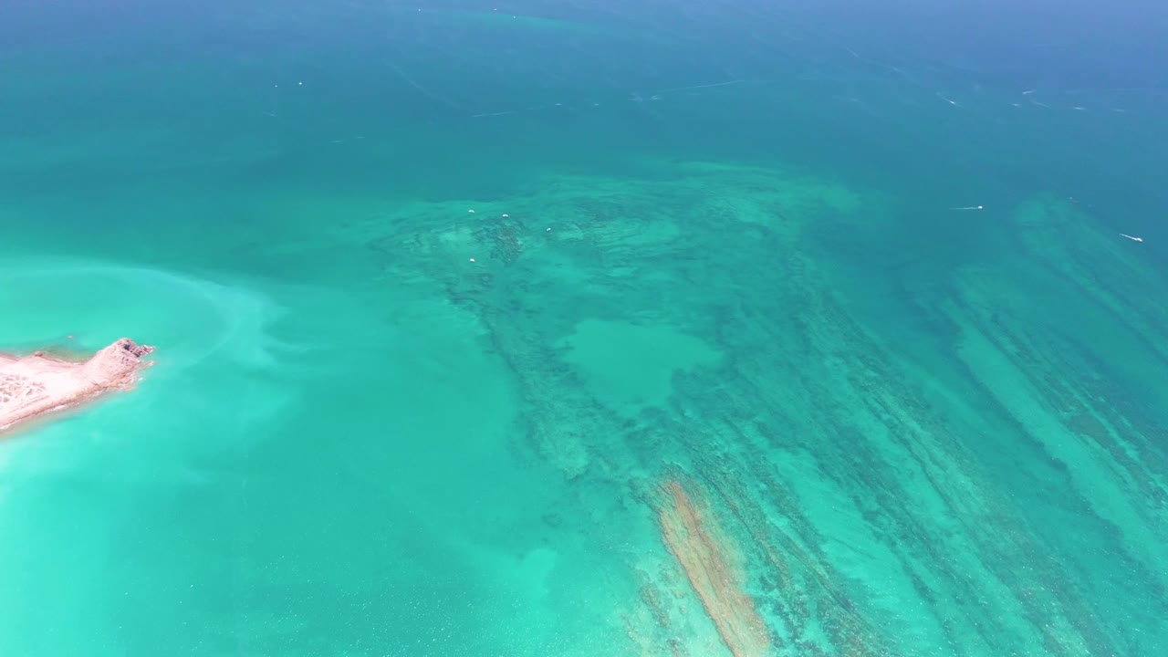 Aerial view in Cabo Pulmo, Mexico, showing a small sandy islet at the left edge, turquoise shallows, darker reef bands, and no visible people.