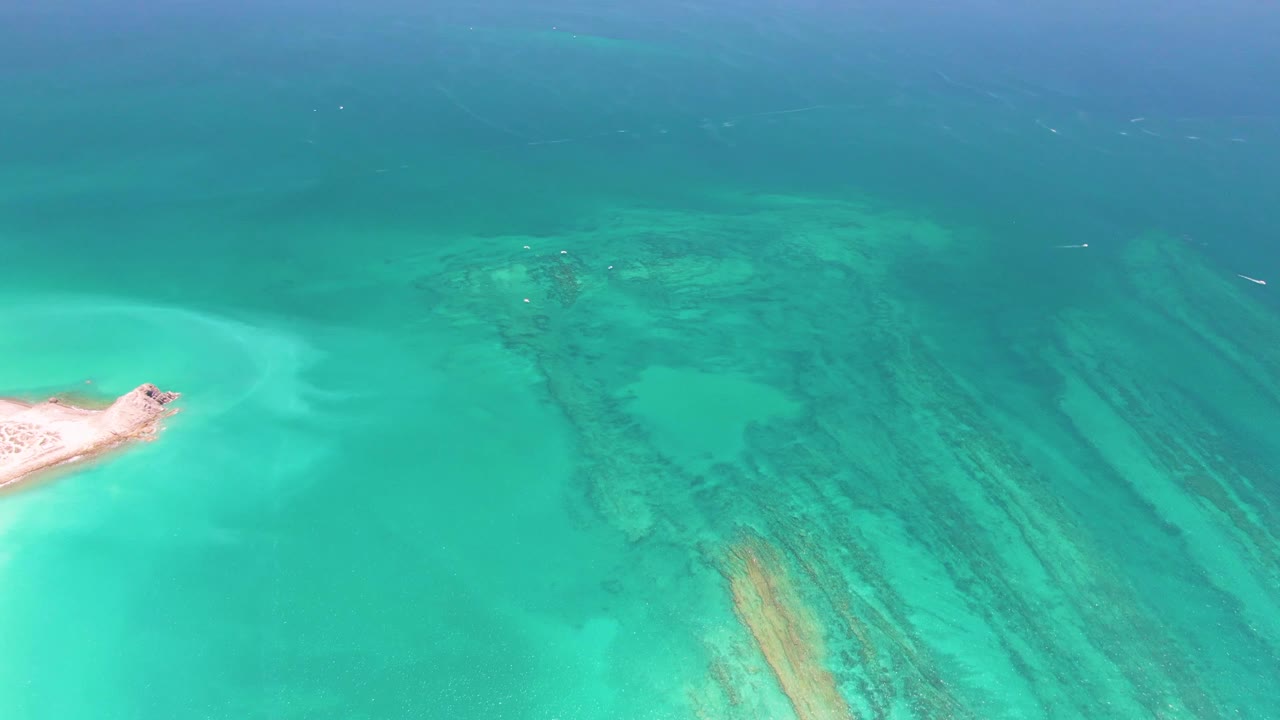 Aerial turquoise shallows with sandy islet at left and reef bands below the surface