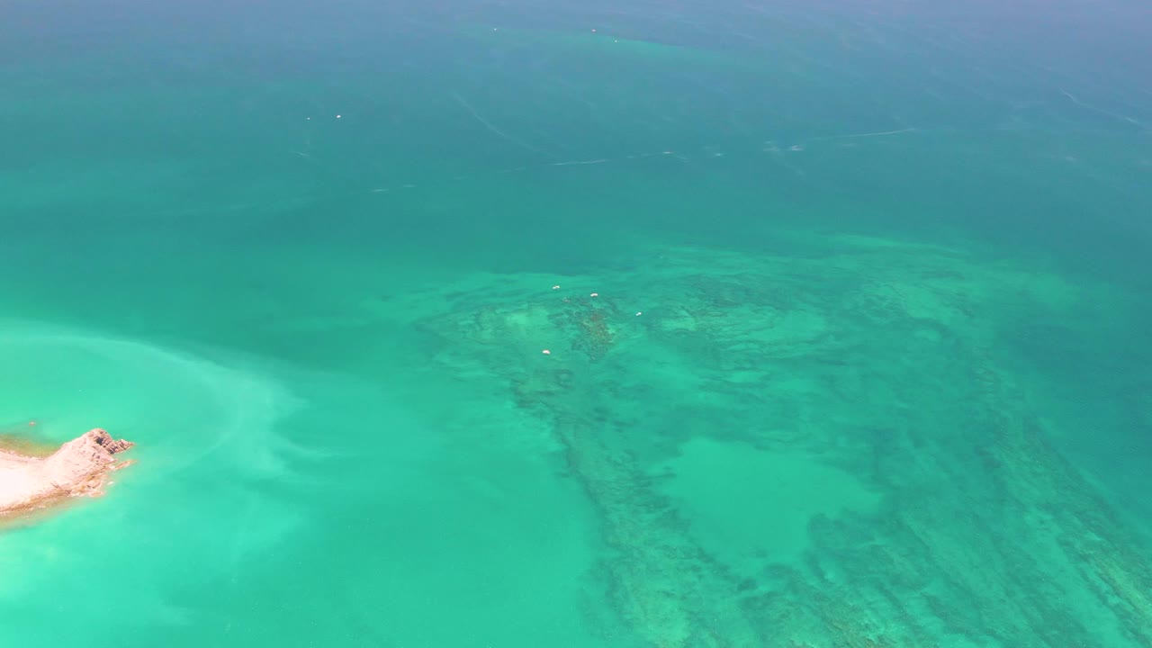 Overhead drone view of clear turquoise water in Cabo Pulmo, Mexico, with pale sand shallows at left, darker seabed patches across the center, and no visible people.
