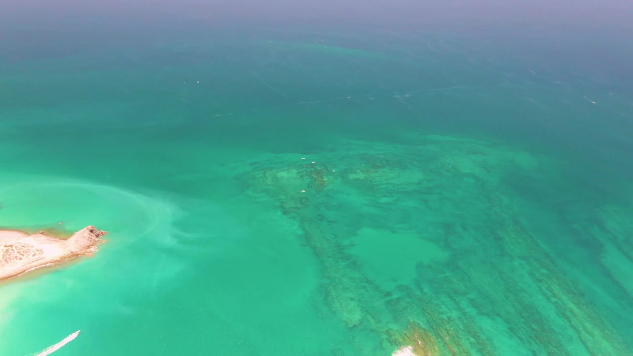 Aerial frame from Cabo Pulmo, Mexico with a small white boat wake beginning near the lower left, a pale sand island at left, reef formations to the right, and no visible people.