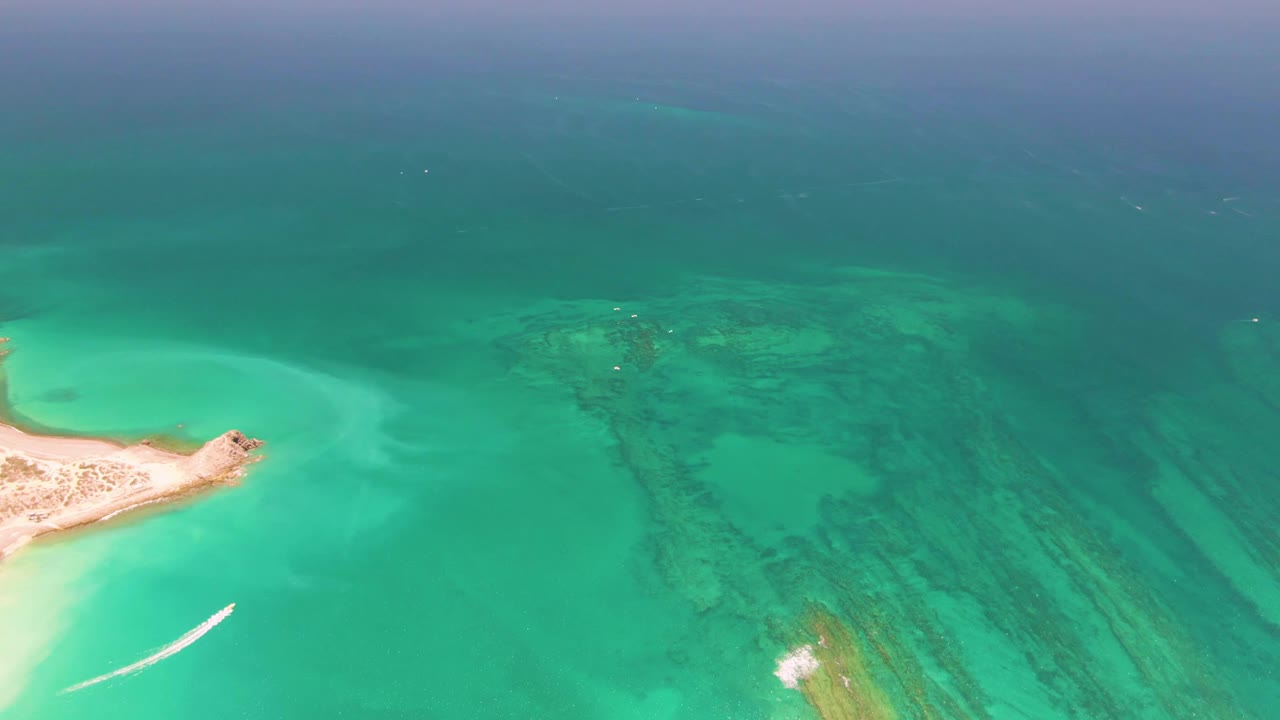 Aerial frame from Cabo Pulmo, Mexico with the sand island more prominent on the left, a white boat wake below it, reef texture across the right side, and no visible people.