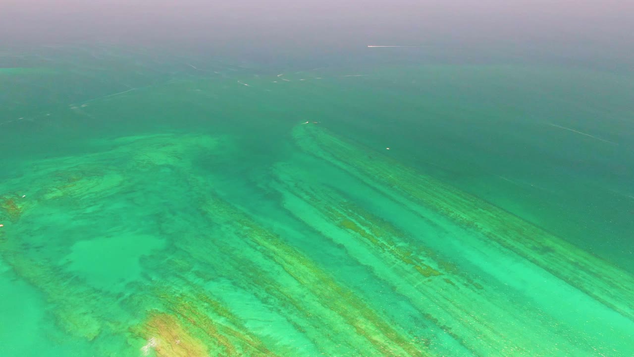 High aerial view of turquoise reef water in Cabo Pulmo, Mexico, with pale sand patches, long submerged reef bands, darker open water near the top, and no visible people.