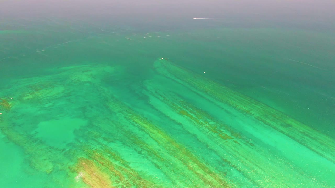 Cabo Pulmo aerial view with broad turquoise shallows, tan reef or rock streaks, pale seabed areas, a hazy darker water line near the horizon, and no visible people.