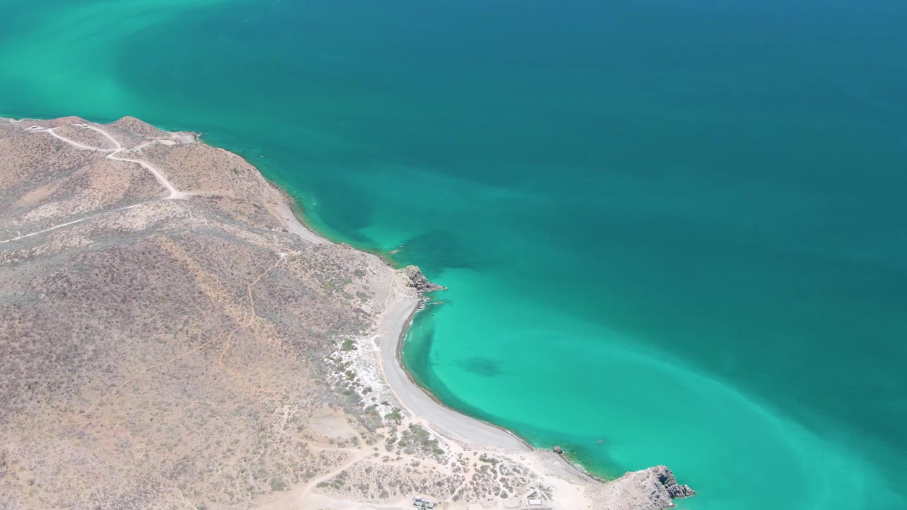 Aerial frame over Cabo Pulmo showing a dry rocky shoreline curving around turquoise water, pale dirt roads on the headland, and no visible people.