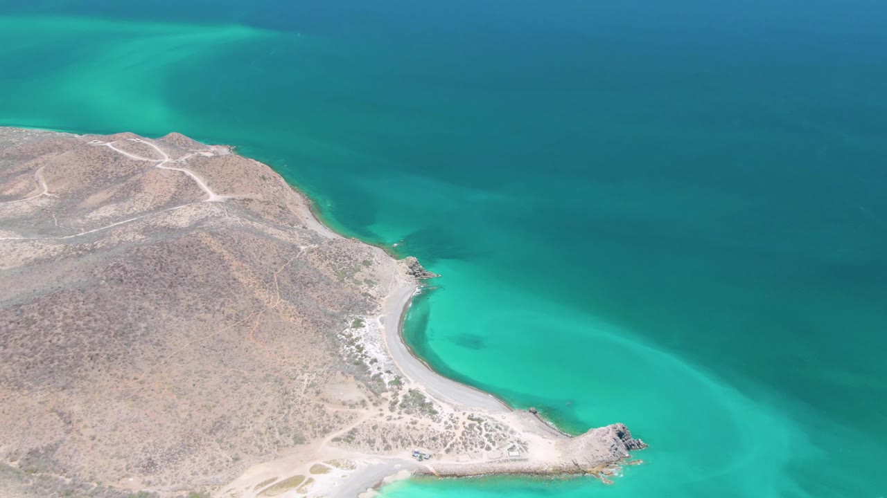 Cabo Pulmo coastline from above with a rocky point at the lower right, light surf along the shore, green-blue shallows, darker offshore water, and no visible people.
