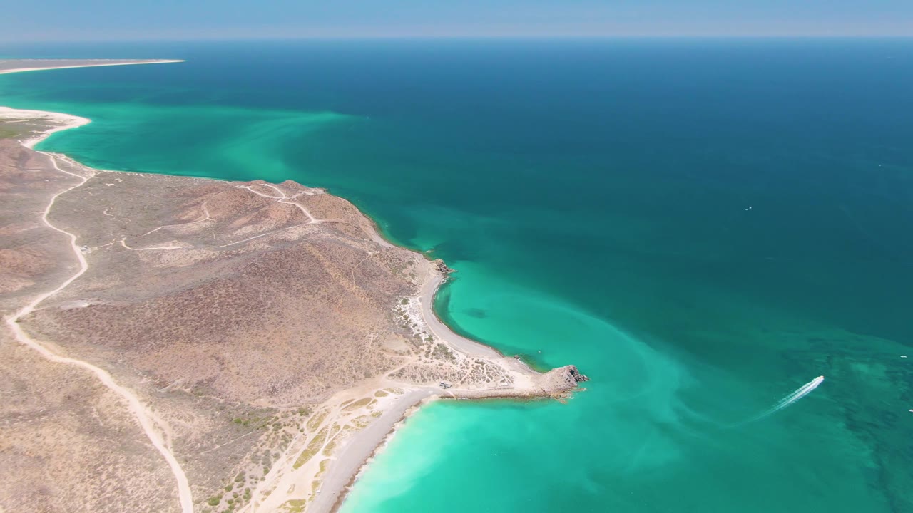 Aerial Cabo Pulmo frame with the peninsula centered lower in view, turquoise water wrapping the shore, a white boat wake to the right, and no visible people.