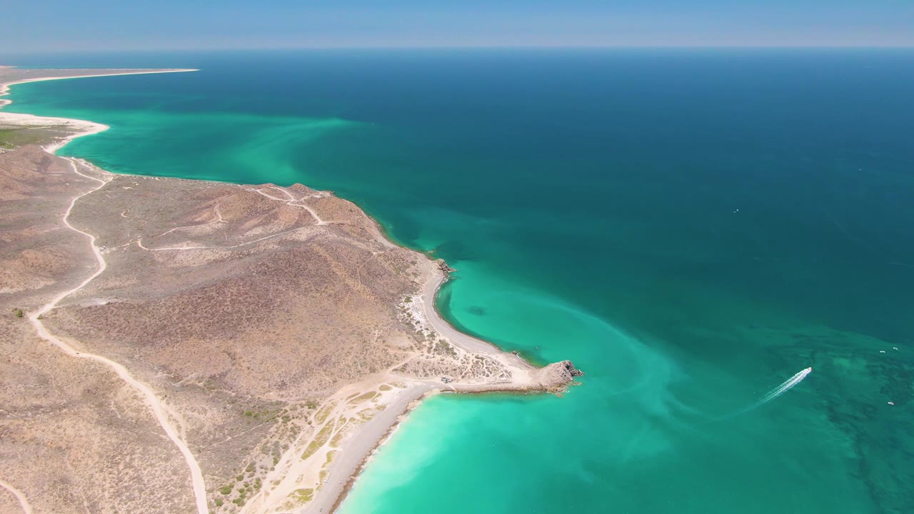Mid-clip aerial frame over Cabo Pulmo with dry peninsula terrain, branching dirt tracks, a curved pale shore, turquoise water, a white wake near the lower right, and no visible people.
