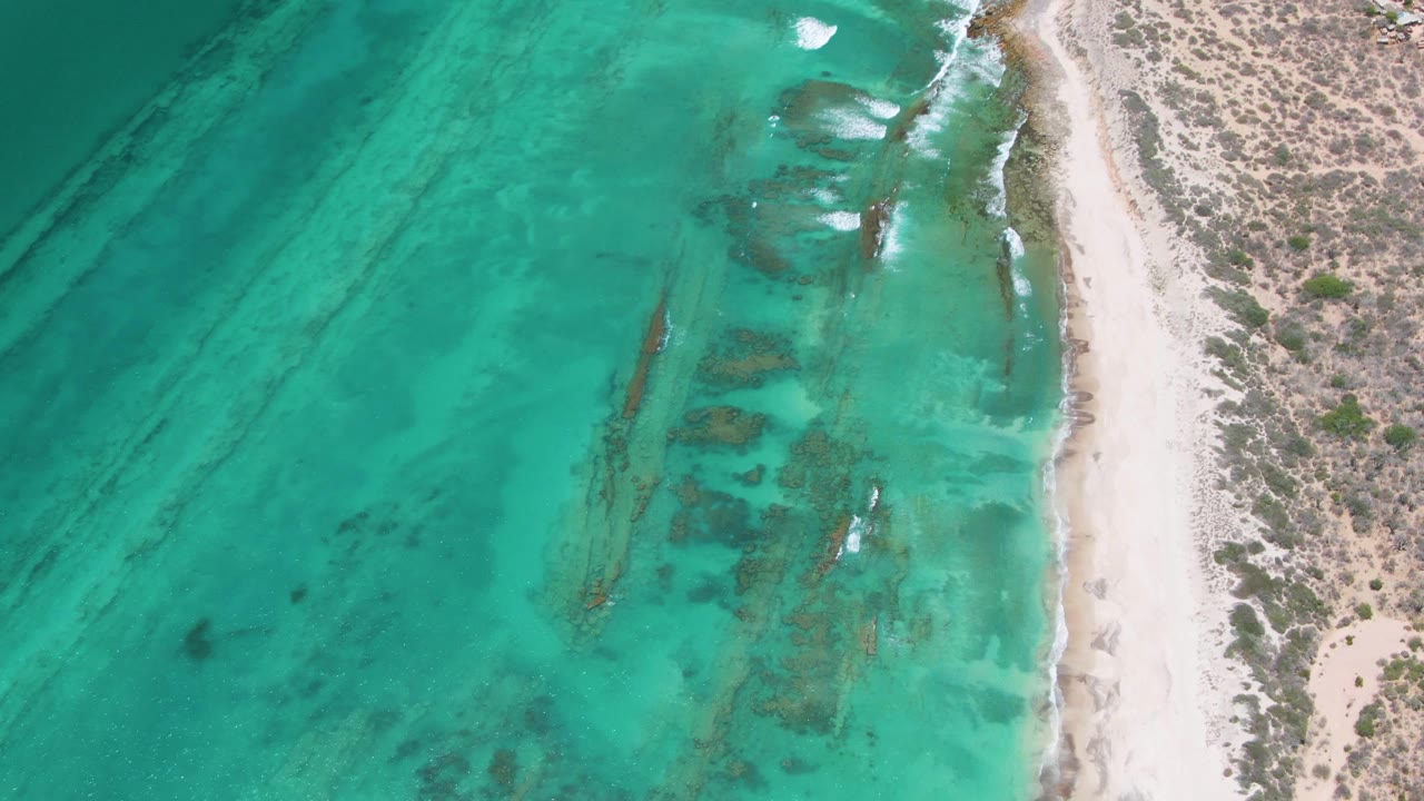 Aerial frame over Cabo Pulmo showing turquoise water, darker submerged lines, a narrow sandy beach, dry dune vegetation, and no visible people at the start of the clip.