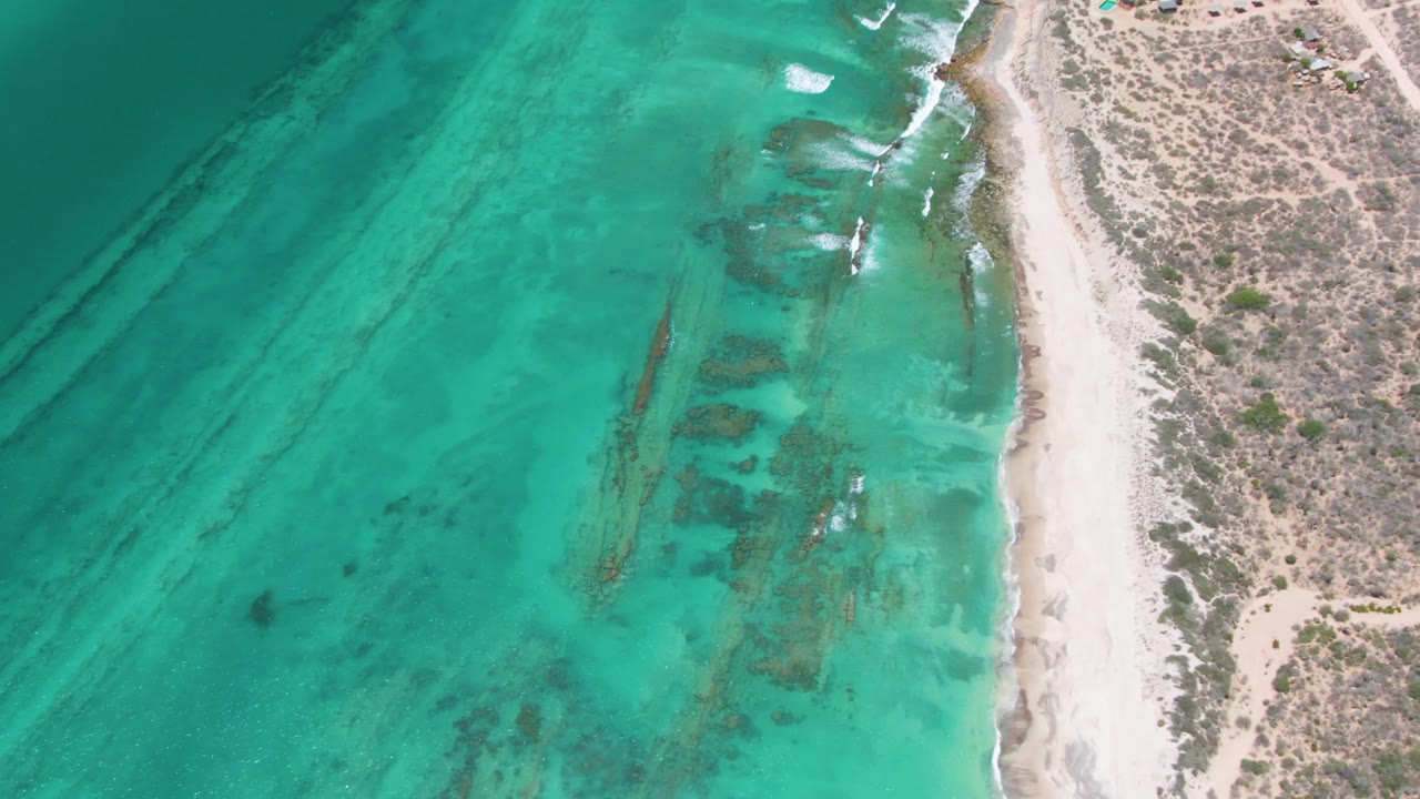 Aerial Cabo Pulmo frame with clear green-blue water, elongated darker underwater streaks, a bright sandy shoreline, scrubby desert ground, and no visible people.
