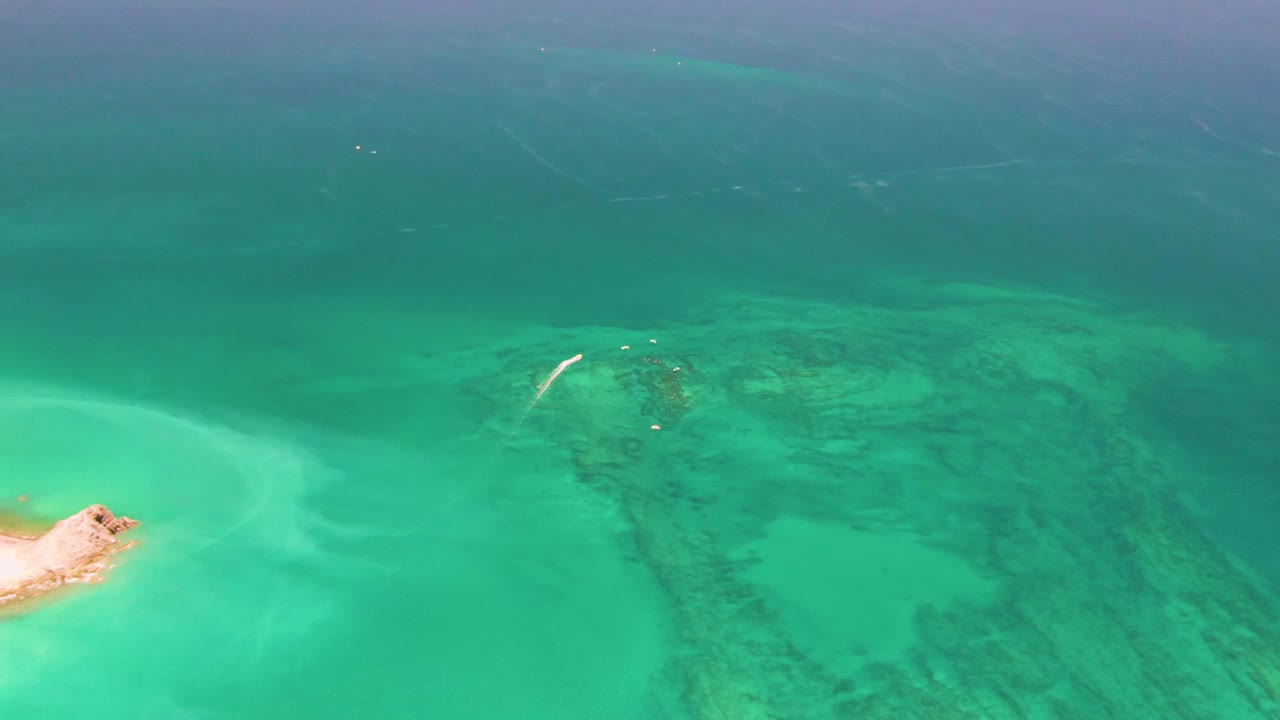 Aerial Cabo Pulmo frame with the sandy islet still visible on the left, clear mint-turquoise water in the foreground, darker coral reef texture across the center, and no visible people.