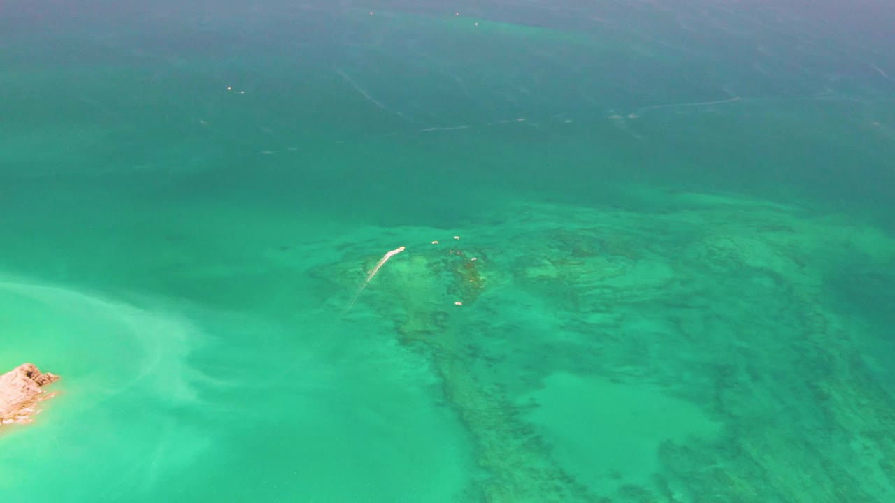 Aerial view over Cabo Pulmo shallows as the sandy islet sits near the left edge, with pale sand swirls, submerged reef patches, teal water, and no visible people.