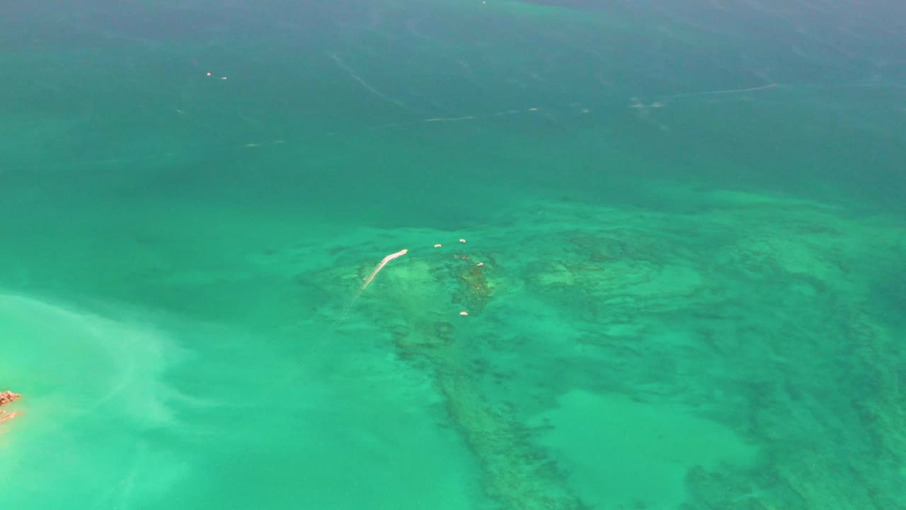 Aerial Cabo Pulmo frame where only a small sliver of sandy islet remains at the lower left, while turquoise water and darker reef formations fill the view with no visible people.