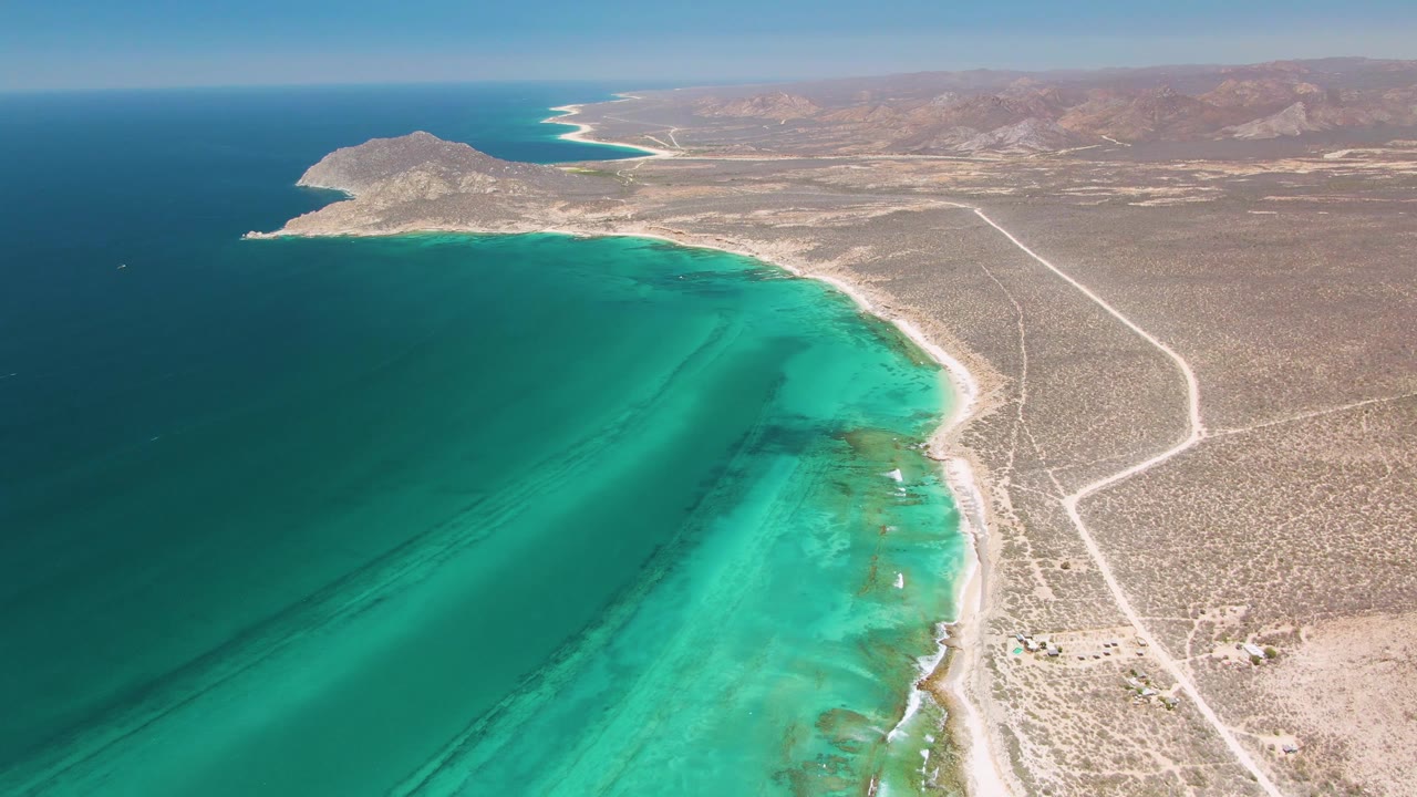 Cabo Pulmo coastline from above, with a broad turquoise band along the shore, dry scrubland roads running inland, low mountains in the distance, and no visible people.
