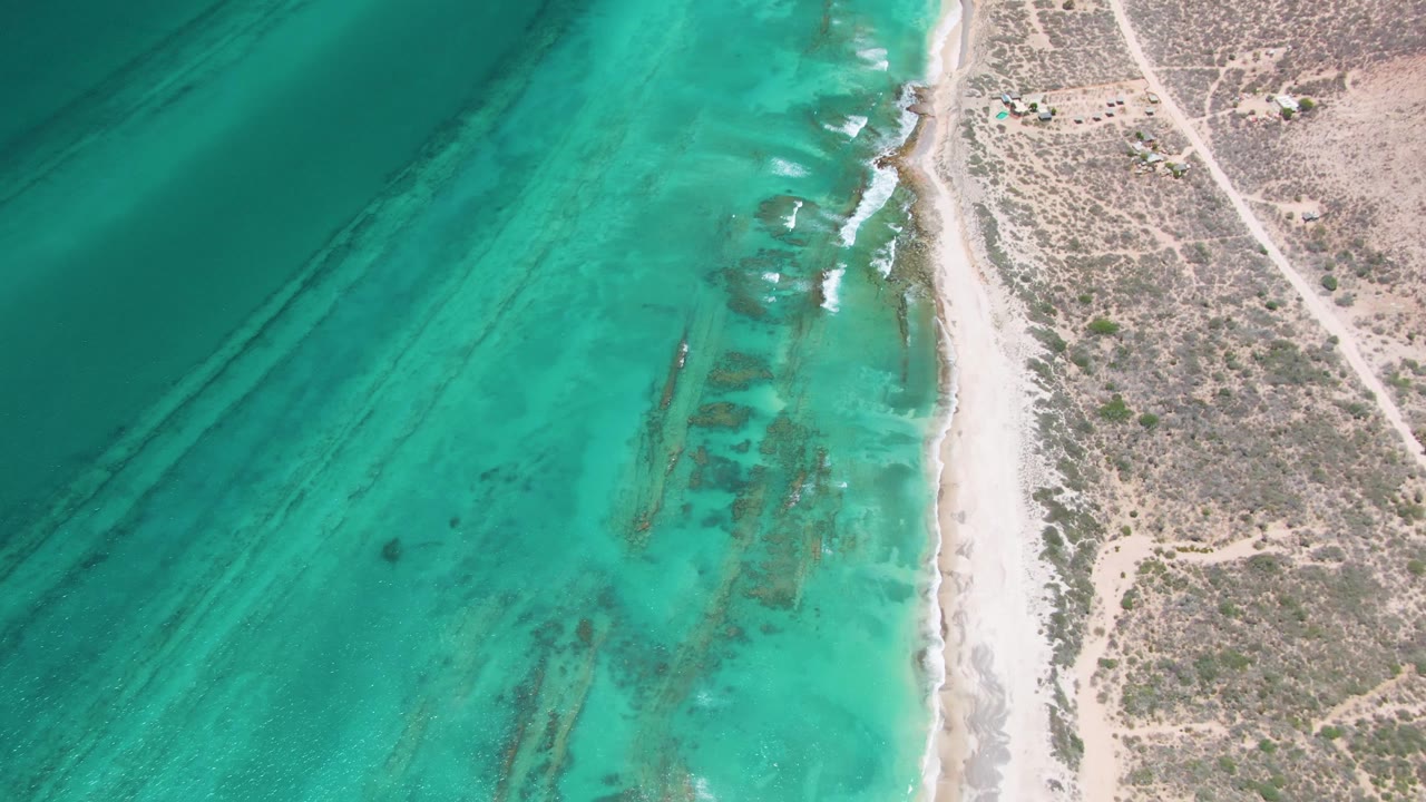 Overhead aerial frame in Cabo Pulmo showing turquoise ocean on the left, a pale sand shoreline near center, dry scrubland on the right, and no visible people.